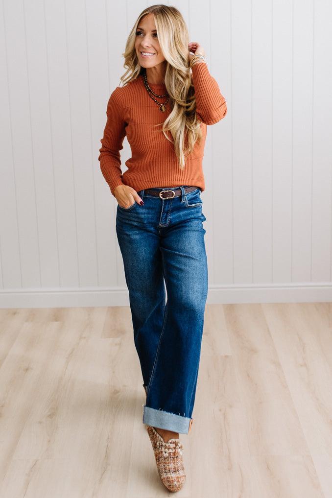 Blonde woman standing in a brightly lit studio with light wood floors and paneled wall behind. Wearing dark pants and a ribbed slim fit long sleeve sweater in rust.
