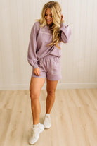 Woman wearing a lavender sweatshirt and shorts set with white sneakers in a room with wooden flooring and a light-colored wall.