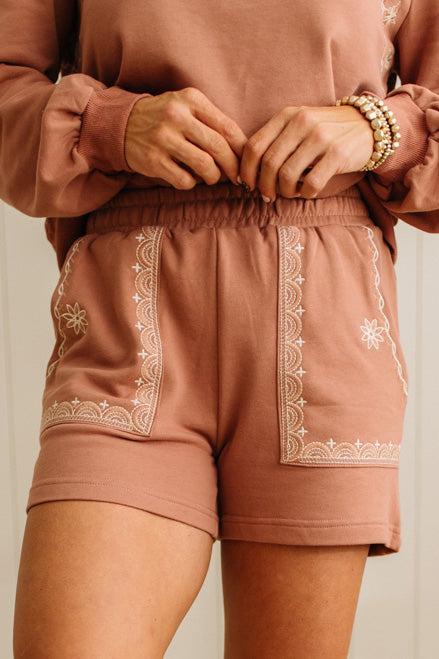 Person wearing a matching brown sweatshirt and shorts set with white embroidery on a neutral background