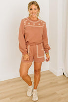 Woman wearing a brown embroidered outfit with white sneakers on a light wooden floor.