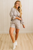Woman wearing a gray sweatshirt and shorts set with white sneakers against a light wooden floor and wall background.