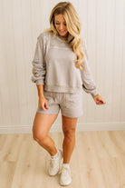 Woman wearing a gray sweatshirt and shorts set with white sneakers in a room with wooden flooring and a white wall.