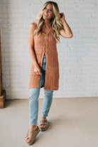 Woman wearing a long brown cardigan over a blue top and jeans, standing against a white brick wall.