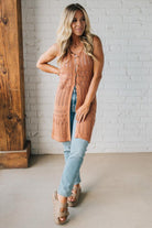 Woman wearing a brown knitted vest and light blue jeans against a white brick wall.