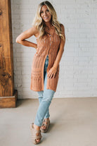 Woman wearing a long brown knitted vest over a light blue top and jeans, standing against a white brick wall.