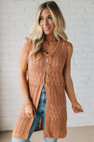 Woman wearing a long brown knitted vest over a blue top against a white brick wall.
