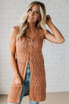 Woman wearing a long brown knitted vest against a white brick wall.
