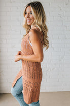 Woman wearing a brown knitted vest and light blue jeans against a white brick wall.