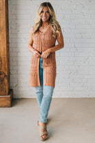 Woman wearing a long brown cardigan over a light blue top and jeans, standing against a white brick wall.