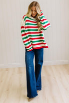 BLONDE WOMAN IN DARK JEANS STANDING IN A BRIGHTLY LIT STUDIO AND WEARING A CHRISTMAS CANDY STRIPE COZY KNIT PULLOVER IN CREAM GREEN AND RED.