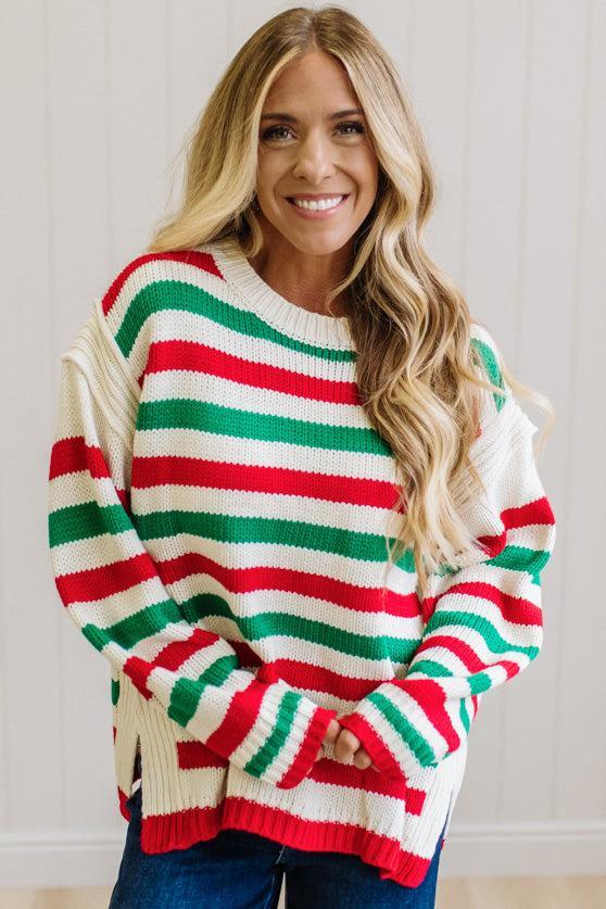 BLONDE WOMAN IN DARK JEANS STANDING IN A BRIGHTLY LIT STUDIO AND WEARING A CHRISTMAS CANDY STRIPE COZY KNIT PULLOVER IN CREAM GREEN AND RED.