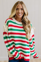 BLONDE WOMAN IN DARK JEANS STANDING IN A BRIGHTLY LIT STUDIO AND WEARING A CHRISTMAS CANDY STRIPE COZY KNIT PULLOVER IN CREAM GREEN AND RED.