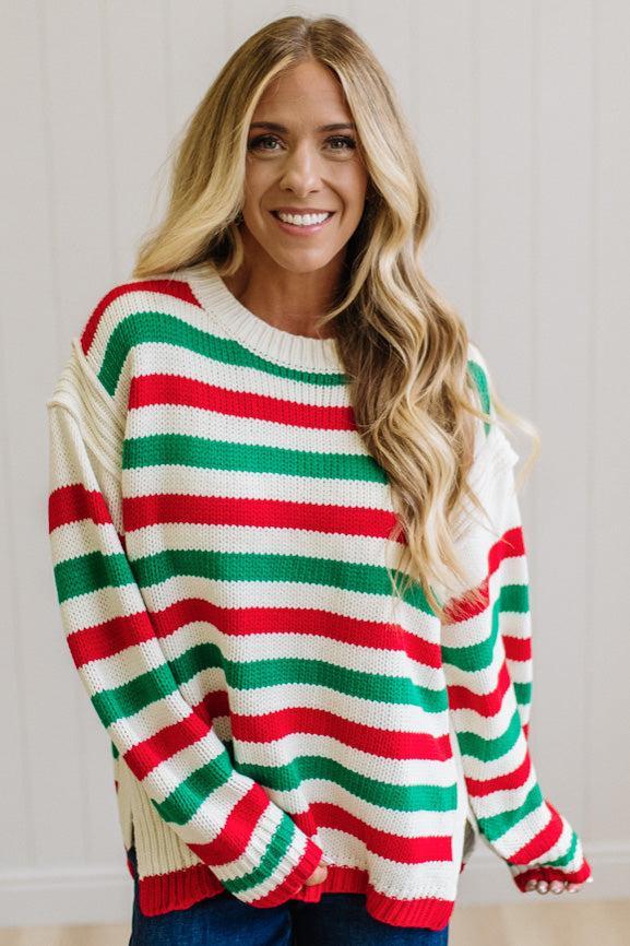 BLONDE WOMAN IN DARK JEANS STANDING IN A BRIGHTLY LIT STUDIO AND WEARING A CHRISTMAS CANDY STRIPE COZY KNIT PULLOVER IN CREAM GREEN AND RED.