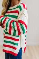 BLONDE WOMAN IN DARK JEANS STANDING IN A BRIGHTLY LIT STUDIO AND WEARING A CHRISTMAS CANDY STRIPE COZY KNIT PULLOVER IN CREAM GREEN AND RED.