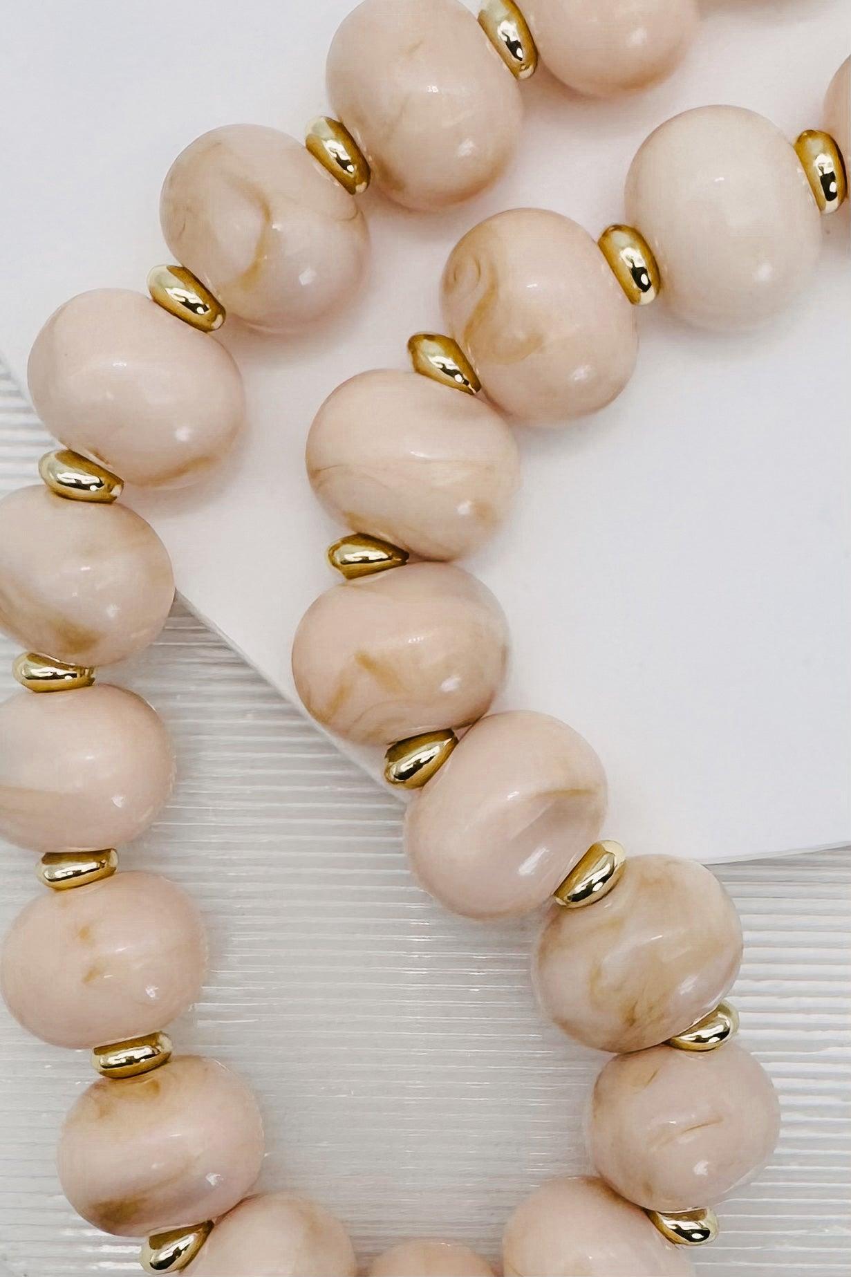 A close-up of a beaded strand with large round pale blush-beige stones separated by small metallic gold spacers, photographed on a white surface.