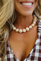 A close-up lifestyle shot of a woman wearing a chunky beaded necklace with cream-colored round beads separated by small gold spacer rings, paired with a brown and white gingham top.