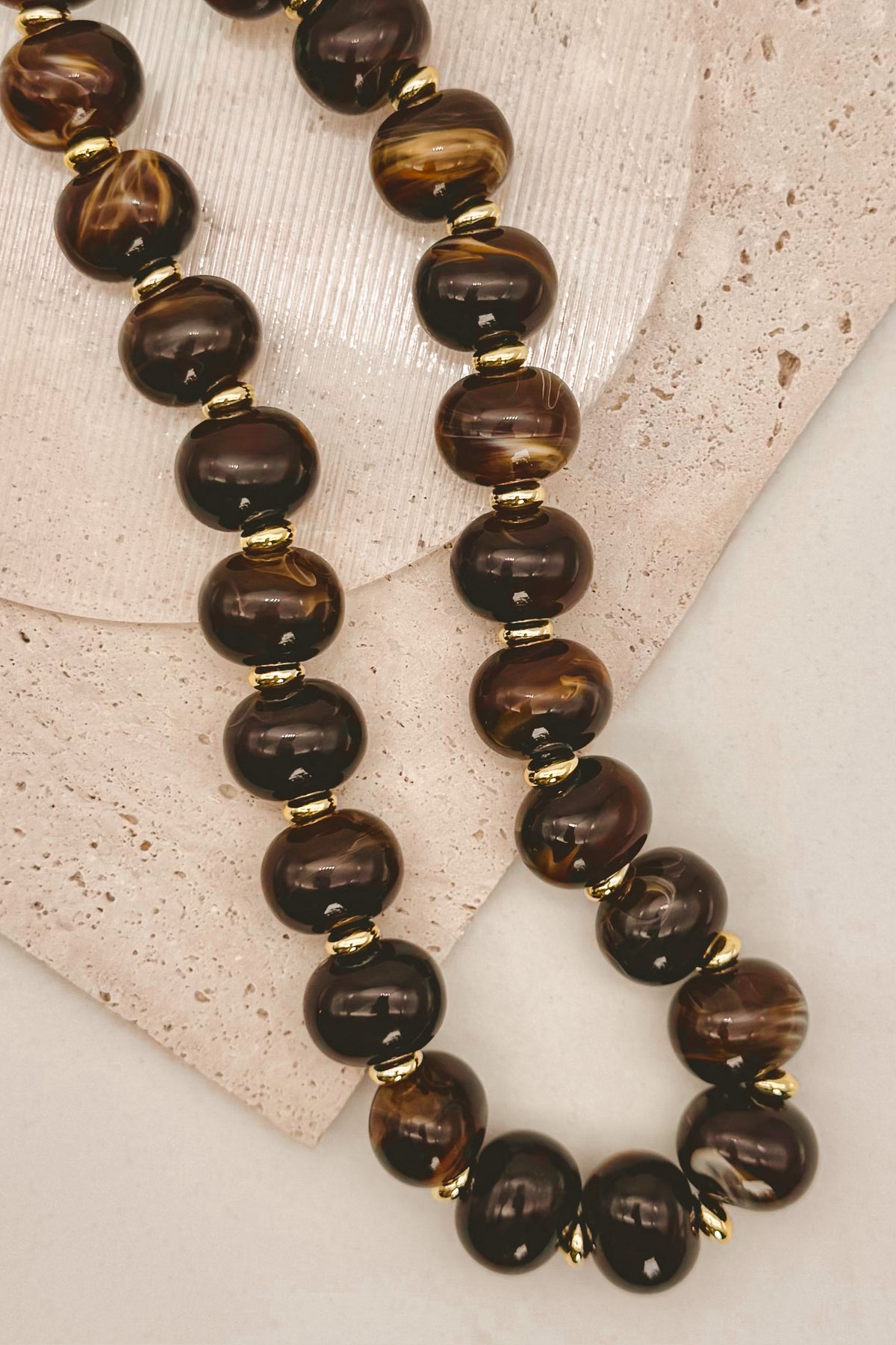 A beaded necklace with large polished brown tiger's eye style beads separated by small gold spacer beads, arranged on a light stone surface.