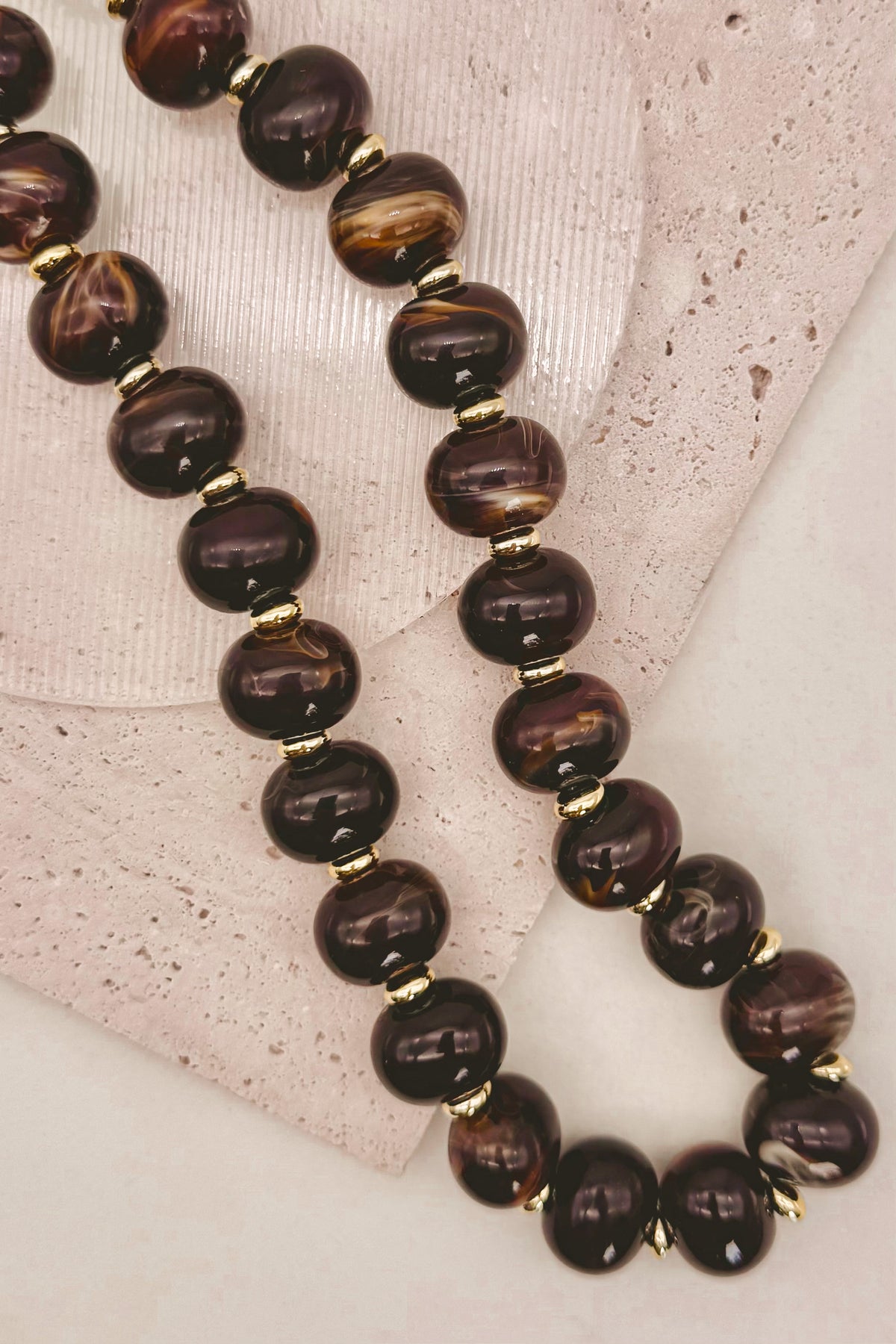 A beaded necklace with glossy dark brown and black marbled beads separated by small gold spacer beads, displayed on a light textured surface.