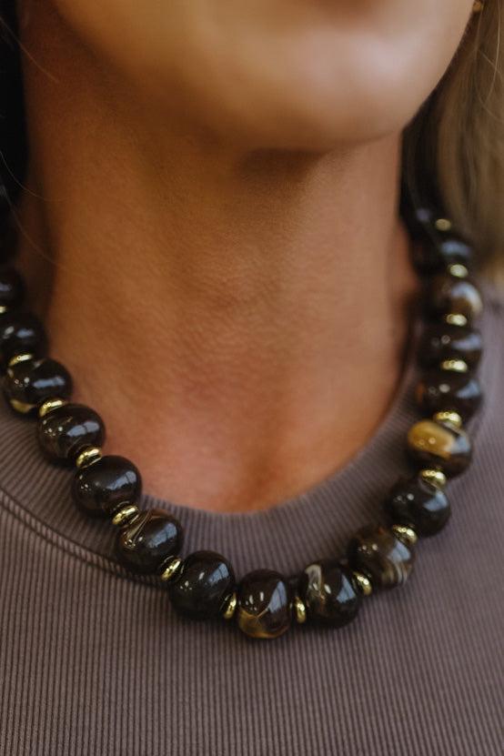 A close-up of a person wearing a beaded necklace made of polished dark brown and black round beads with small gold spacer beads, paired with a taupe ribbed top.