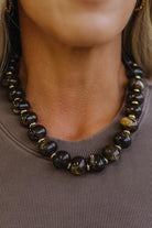 A close-up of a woman wearing a chunky beaded necklace made of dark brown and black polished stone beads with small gold spacer accents, paired with a taupe ribbed top.