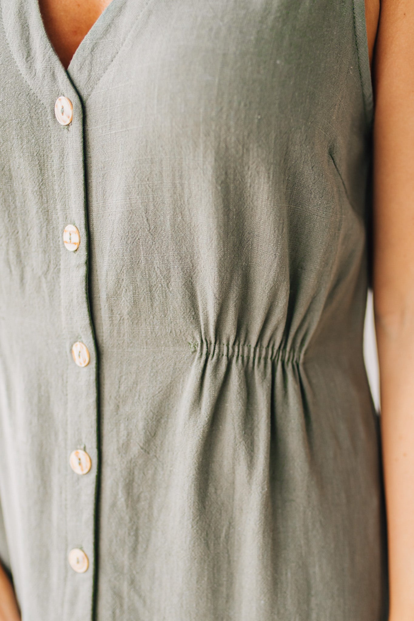 BLONDE WOMAN WEARING A LIGHT OLIVE LINEN BLEND V NECK BUTTON DOWN SLEEVELESS DRESS.