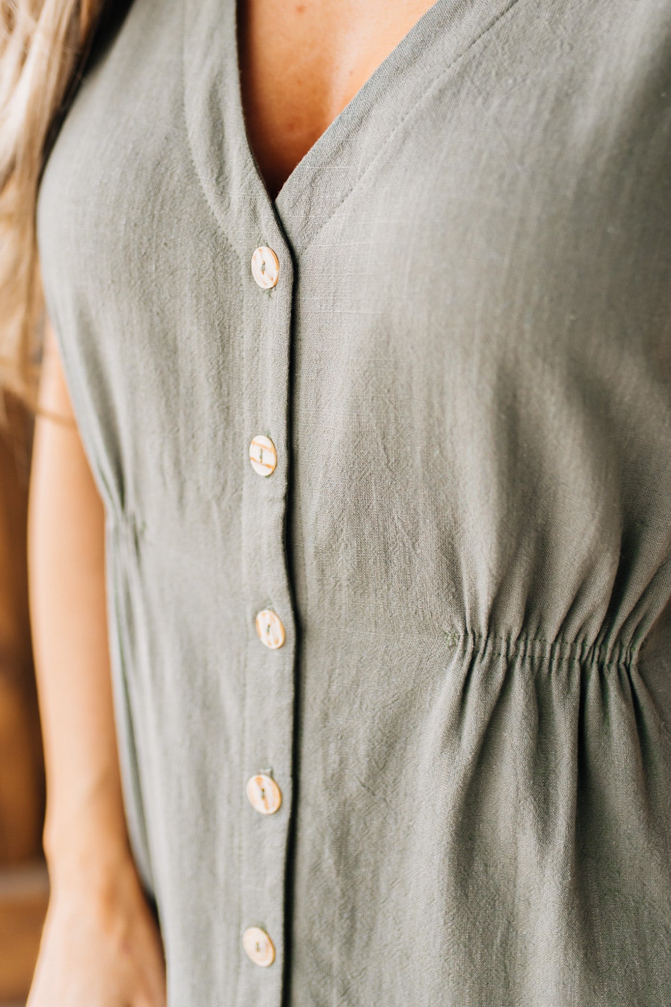 BLONDE WOMAN WEARING A LIGHT OLIVE LINEN BLEND V NECK BUTTON DOWN SLEEVELESS DRESS.