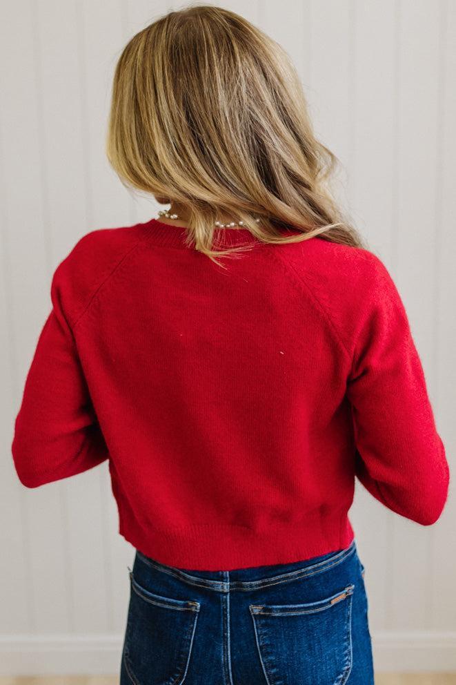 BLONDE WOMAN IN A LIGHT STUDIO WEARING DARK JEANS AND A LIGHT TOP WITH A RED CARDIGAN OVER TOP THAT HAS LARGE LACE BOWS ON THE SIDES.