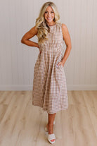 Woman wearing a checkered dress standing in a room with wooden flooring and a light-colored wall.