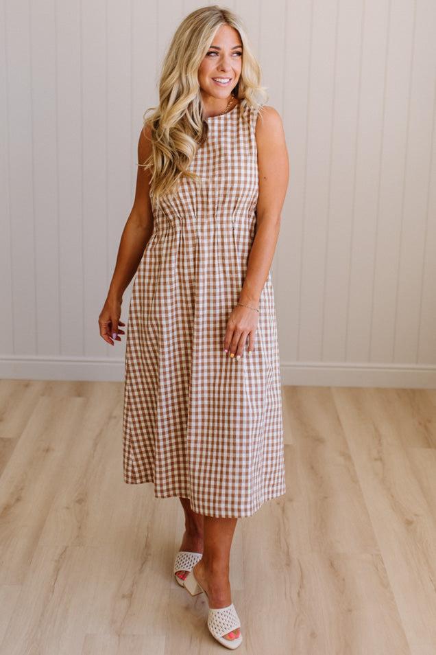 Woman wearing a checkered dress standing in a room with wooden flooring and a white paneled wall.