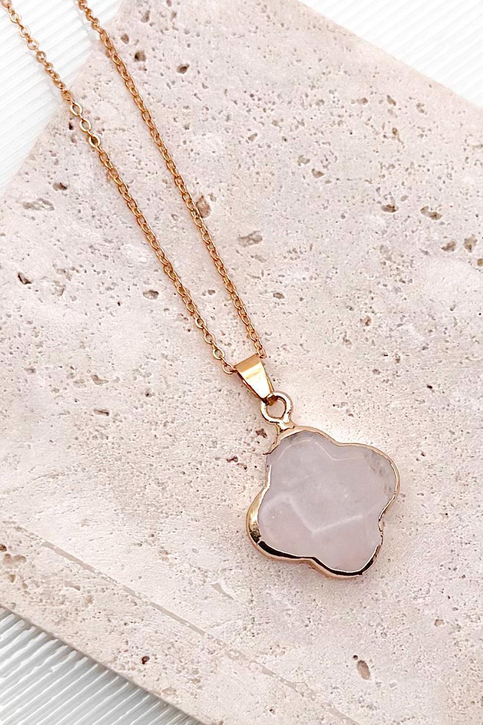 A gold necklace with a wavy clover-shaped pendant made of grey lazuli stone, displayed against a white background with vertical gray ribbed lines.