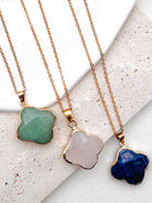 Three gold-tone chain necklaces with irregularly shaped stone pendants in green, pale pink, and deep blue, arranged on a light textured stone surface.