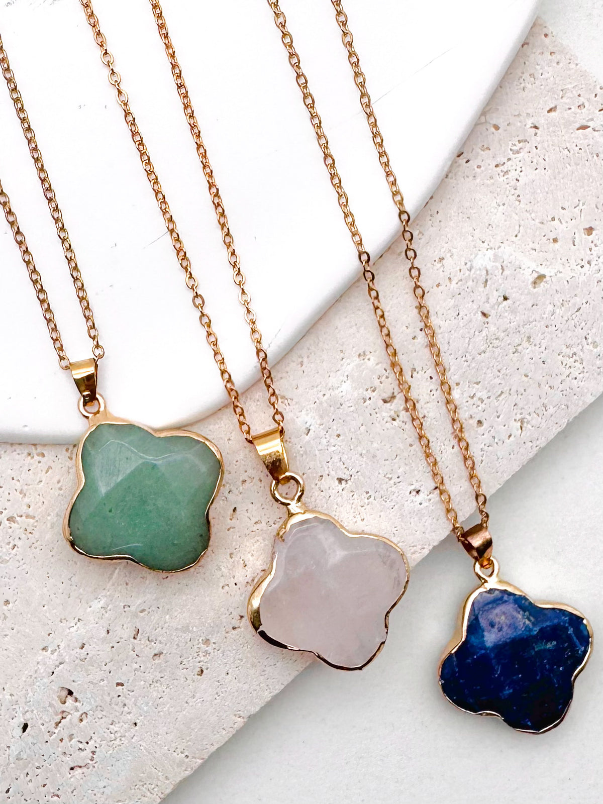 Three gold-tone chain necklaces with irregularly shaped stone pendants in green, pale pink, and deep blue, arranged on a light textured stone surface.