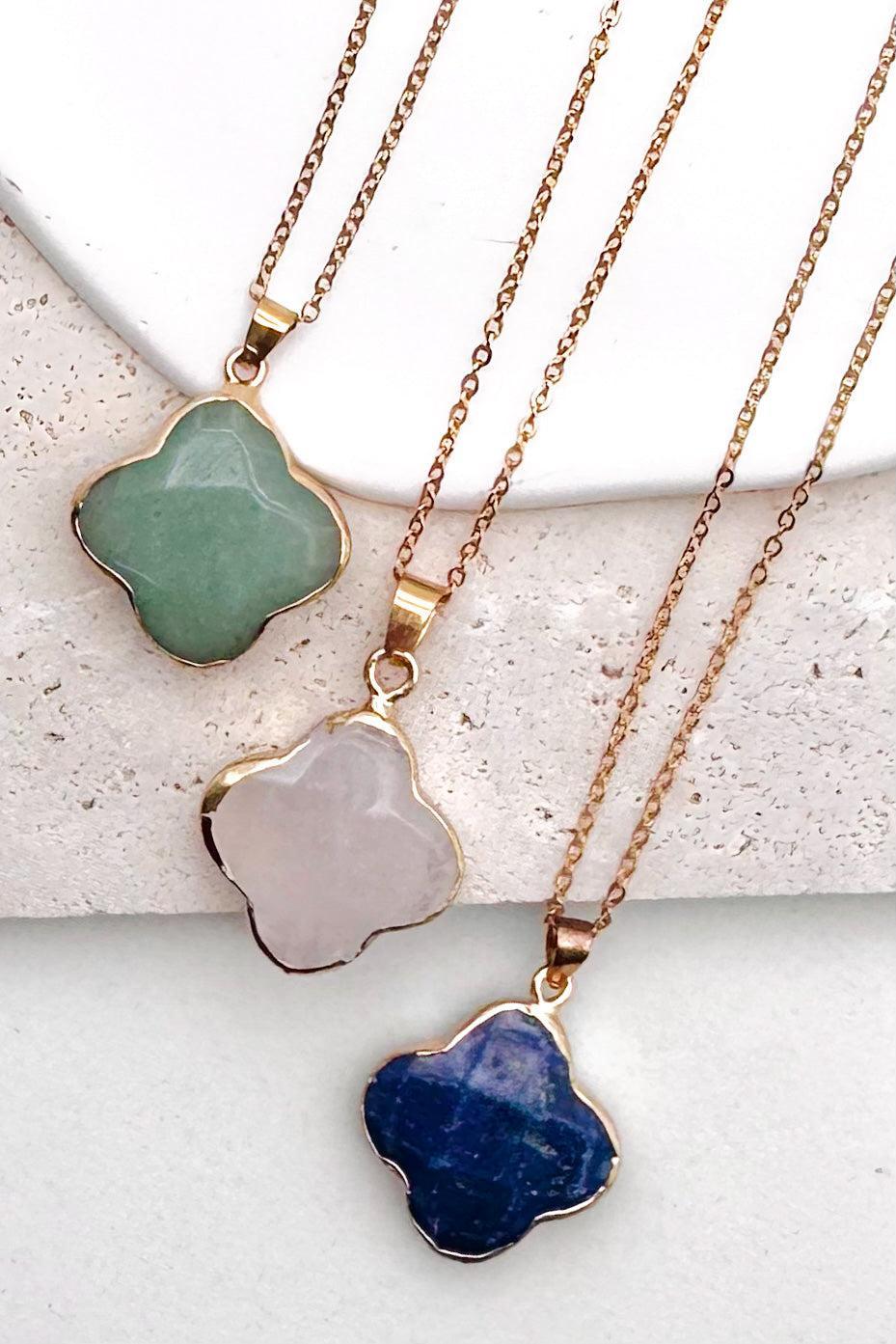 Three gold-tone chain necklaces with irregular clover-shaped stone pendants are arranged diagonally on a textured white surface. The pendants are green, pale pink, and deep blue.