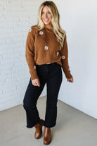Woman wearing a brown floral sweater, black pants, and brown boots against a white brick wall.