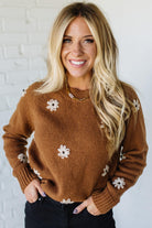 Woman wearing a brown sweater with white floral patterns against a white brick wall.