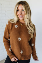 Woman wearing a brown sweater with white floral patterns against a white brick wall.