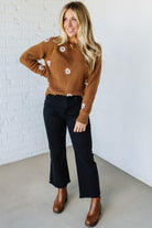Woman wearing a brown sweater with white floral patterns, black pants, and brown boots against a white brick wall.