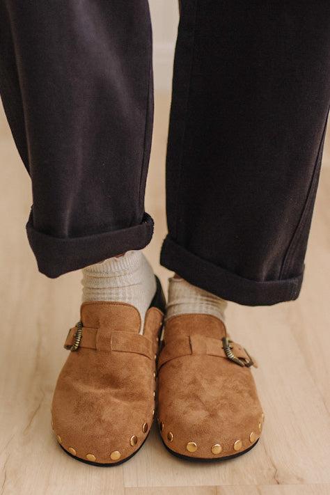 A close-up of a person's feet wearing brown suede clogs with gold-tone studs and buckles, paired with white socks and dark charcoal pants on a light wood floor.
