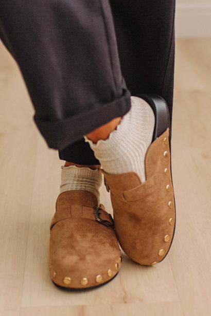 A close-up of a person wearing brown suede slip-on mules with gold-tone studs, paired with white ribbed socks and dark pants, standing on a light wood floor.