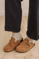 A person wearing brown suede clogs with gold-tone studs and buckles, paired with white socks and dark pants, standing on a light wood floor.