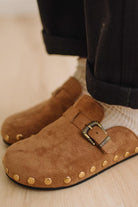 A close-up of a person wearing brown suede slip-on clogs with brass-tone buckles and round metal studs, paired with white socks and dark pants on a light wood floor.