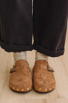 A close-up of a person's feet wearing tan suede clogs with gold-tone studs and buckles, paired with light beige socks and dark cuffed trousers on a light wood floor.