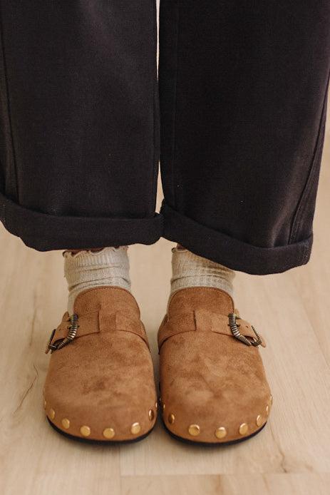A close-up of a person's feet wearing tan suede clogs with gold-tone studs and buckles, paired with light beige socks and dark cuffed trousers on a light wood floor.