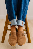 A close-up of a person wearing brown suede slip-on clogs with brass-tone buckles and round metal studs, paired with white socks and dark pants on a light wood floor.
