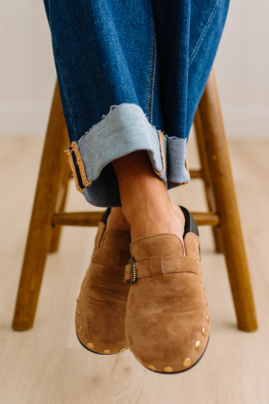 A close-up of a person wearing brown suede slip-on clogs with brass-tone buckles and round metal studs, paired with white socks and dark pants on a light wood floor.