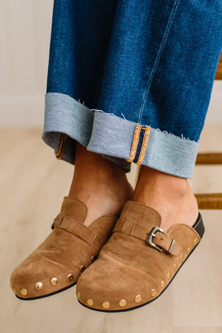 A close-up of a person wearing brown suede slip-on clogs with brass-tone buckles and round metal studs, paired with white socks and dark pants on a light wood floor.