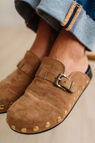 A close-up of a person wearing brown suede slip-on clogs with brass-tone buckles and round metal studs, paired with white socks and dark pants on a light wood floor.