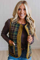 A smiling blonde woman wearing a green and black plaid quilted vest with olive green trim over a dark brown long-sleeve top and blue jeans, standing against a light neutral wall.