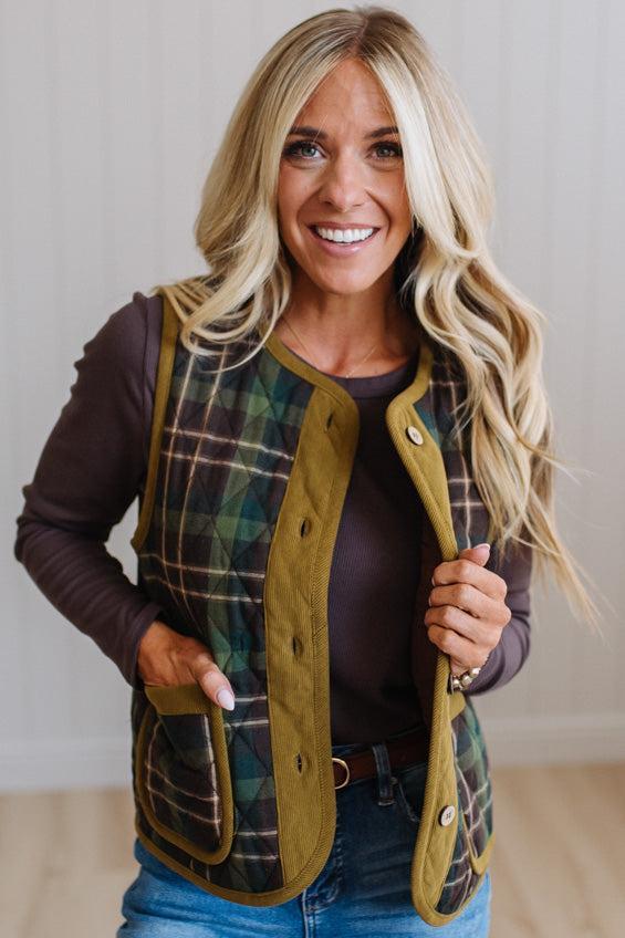 A smiling blonde woman wearing a green and black plaid quilted vest with olive green trim over a dark brown long-sleeve top and blue jeans, standing against a light neutral wall.