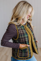 A woman wearing a green and black plaid quilted vest with olive green trim over a dark plum long-sleeve shirt and blue jeans, shown in a side profile against a neutral indoor background.